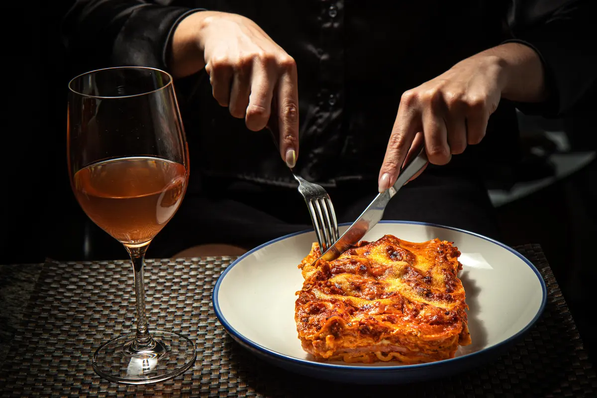Mãos de um convidado a cortar lasanha, com um copo de vinho rosé.