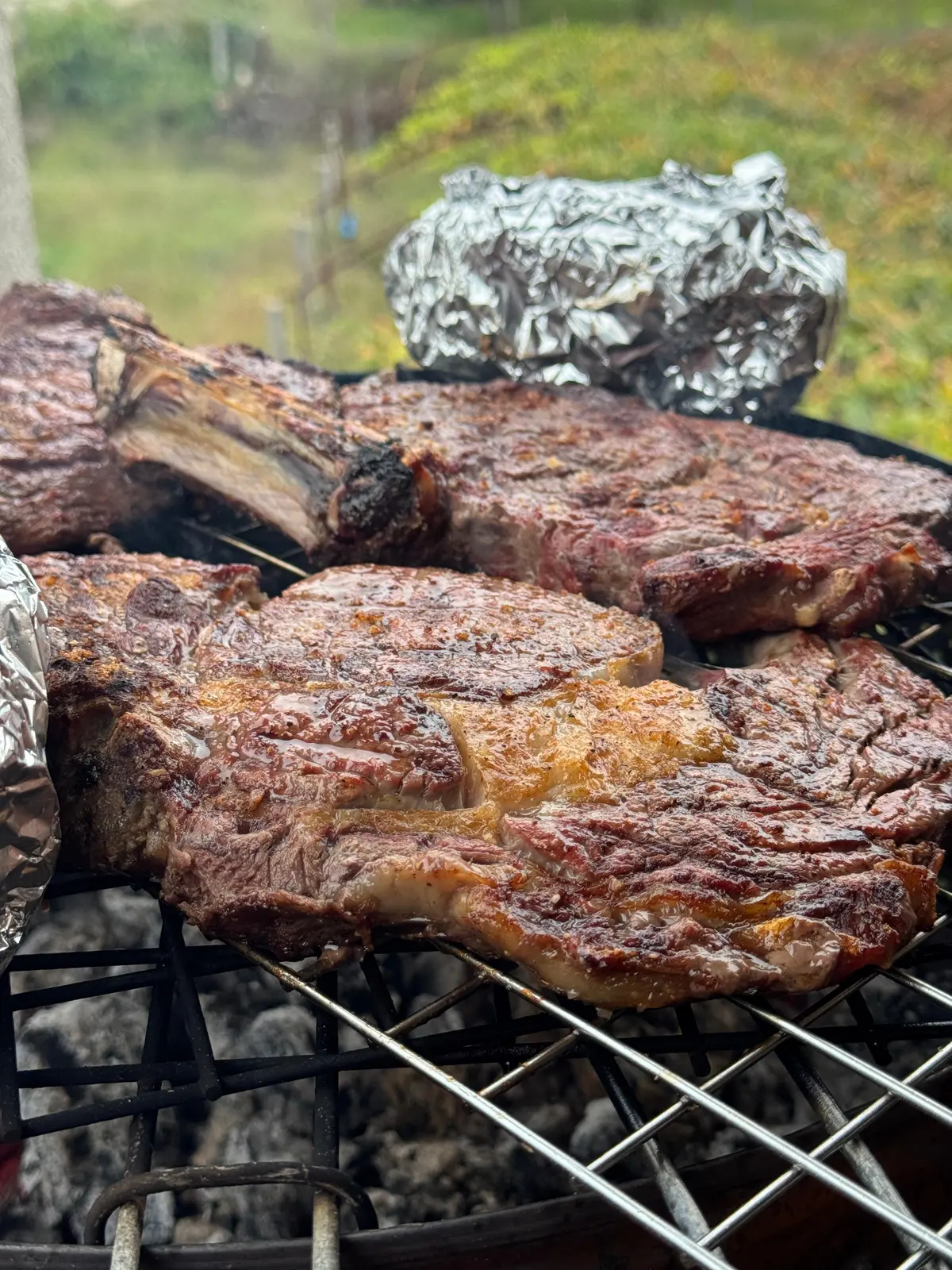 Bifes grossos a grelhar sobre brasas, ao ar livre.