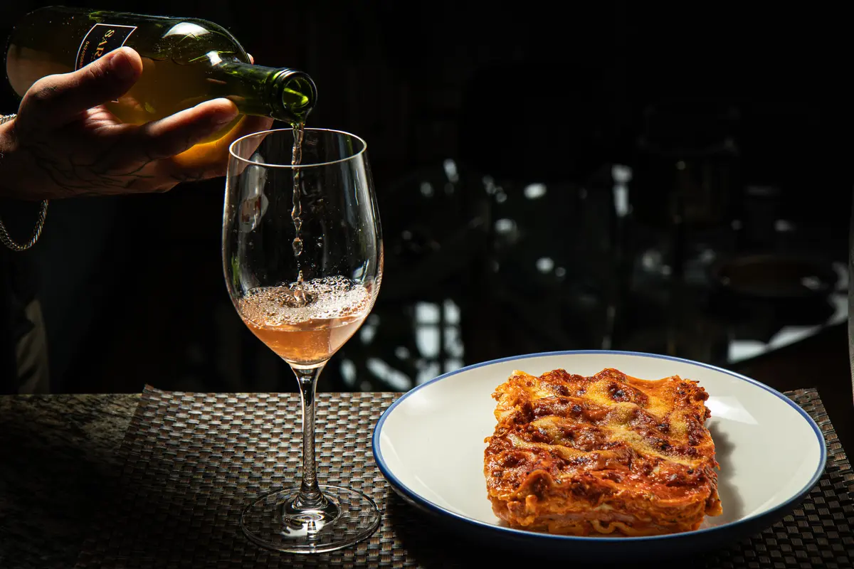 Vinho a ser servido num copo, ao lado de um prato de lasanha.